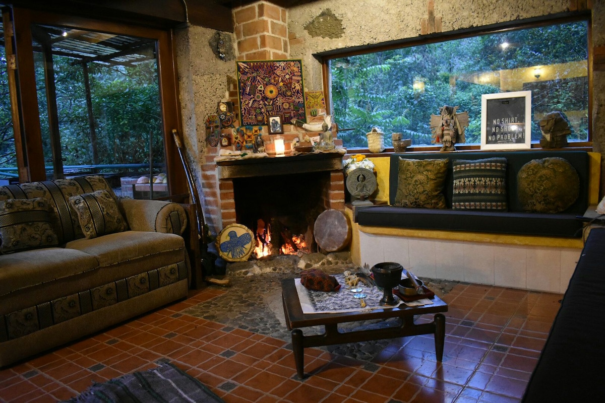 A welcoming living space features a cozy seating area with a patterned couch and cushions. A stone fireplace occupies one corner, with a warm glow emanating from the flames. Large windows provide views of the surrounding nature, enhancing the tranquil atmosphere.