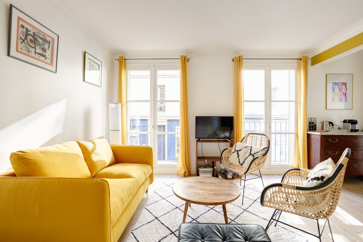 A bright living area features a yellow sofa paired with two woven chairs, complemented by a wooden coffee table. Natural light flows through large windows adorned with yellow curtains, illuminating the space. A TV is positioned on a dark wood console against the wall.