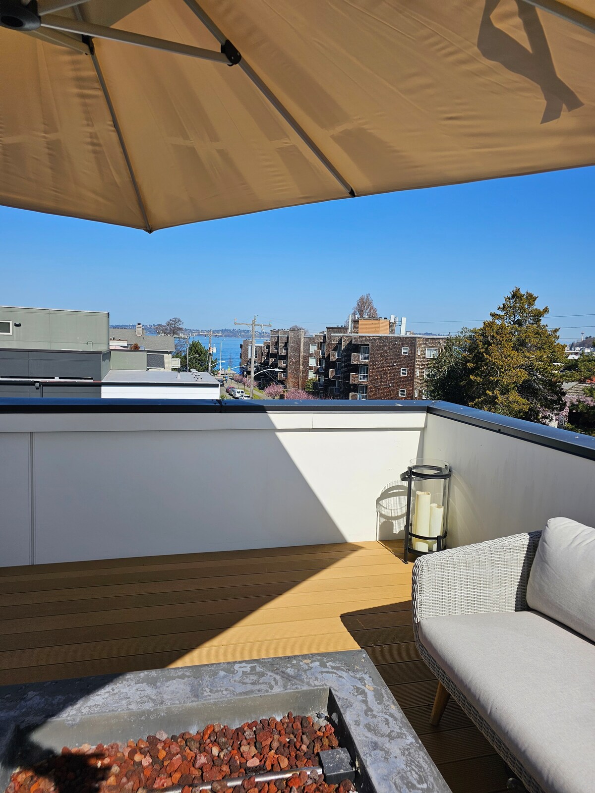 A sunny roof deck is showcased, featuring a comfortable seating area with a light-colored chair. A firepit is at the center, surrounded by decorative stones. In the background, views of the water and neighboring buildings are visible under a clear blue sky.