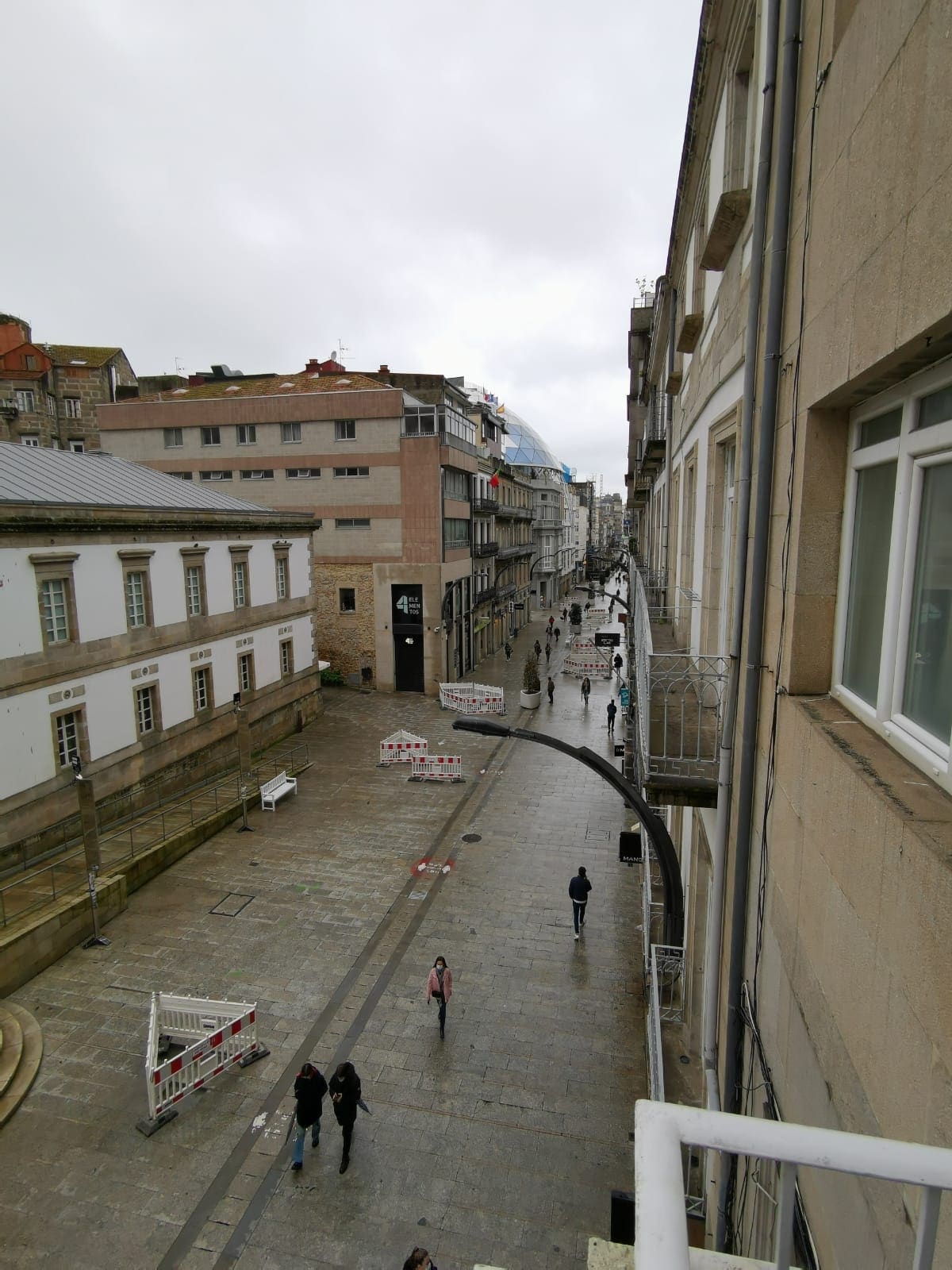 A wide view of a pedestrian street captures a mix of modern and traditional buildings. Soft grey skies create a muted atmosphere. Paved pathways extend along either side, with people walking leisurely. Barriers are positioned intermittently, indicating areas of activity or construction.