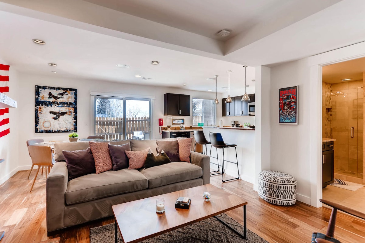 A spacious living area features a light gray sofa adorned with decorative pillows, accompanied by a wooden coffee table. Nearby, a modern kitchen with bar seating is visible, along with a dining table. Natural light fills the room through large windows, while recessed lighting complements the bright ambiance.