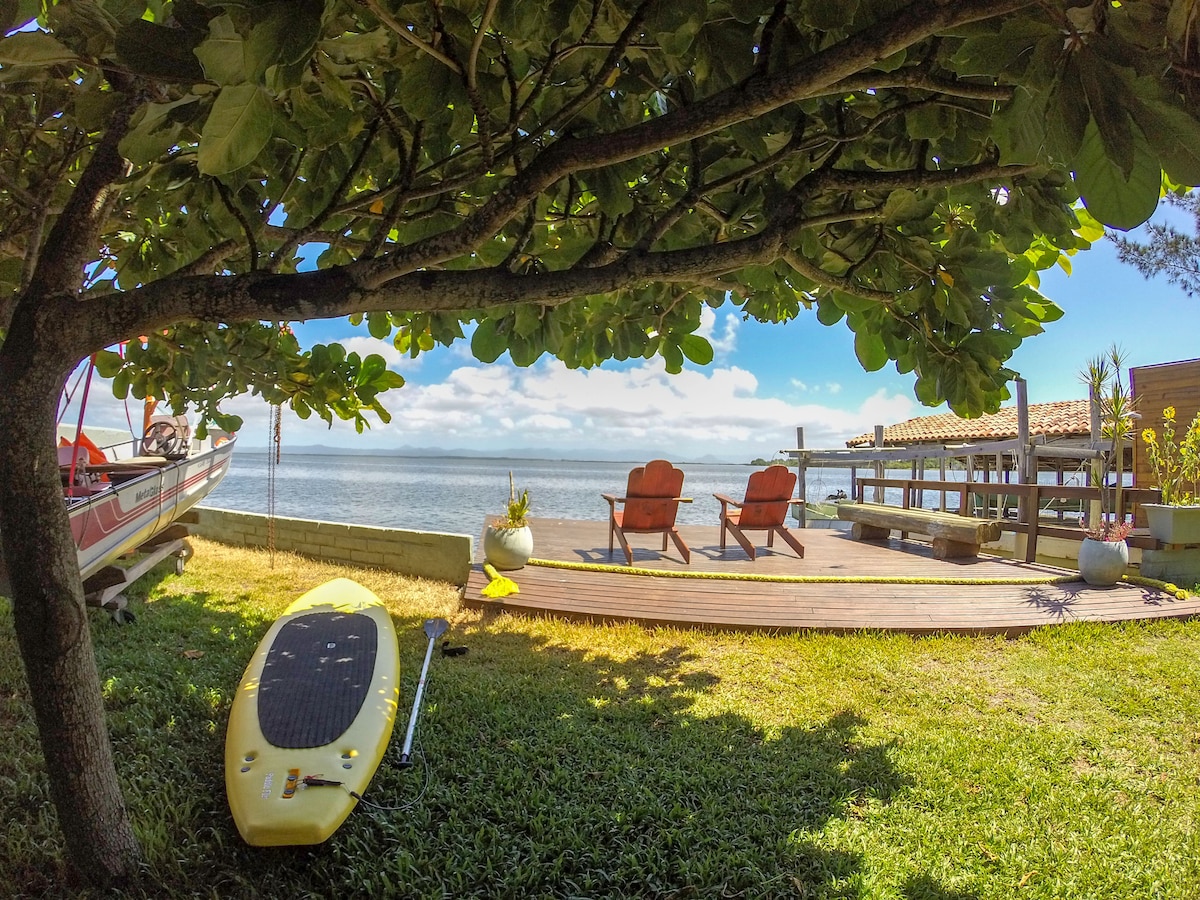 A serene outdoor space overlooking the lagoon features two wooden chairs positioned on a deck. A vibrant green tree provides shade, while a yellow paddleboard rests on the grass nearby. The tranquil water reflects the sky, offering a peaceful atmosphere.