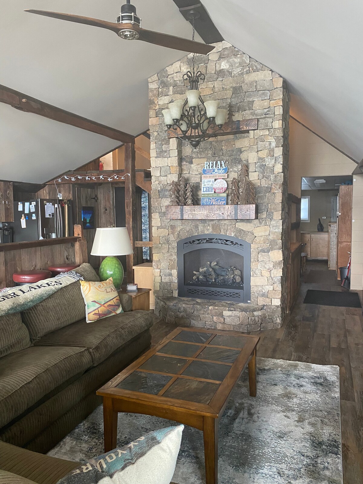 A living area features a stone fireplace as the focal point, surrounded by wooden beams and high ceilings. A comfortable sofa is positioned near a coffee table, with a lamp providing soft illumination. A rug adds texture to the floor, enhancing the cozy atmosphere.
