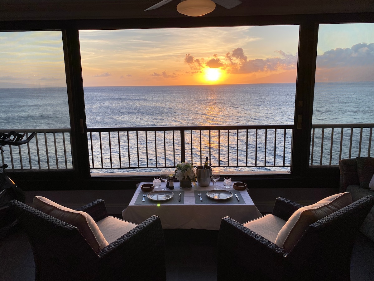 A dining setup is featured on an oceanview lanai, with a table elegantly set for two. Chairs offer a comfortable seating area, and the stunning sunset casts a warm glow over the calm waters, creating a serene atmosphere for meals.