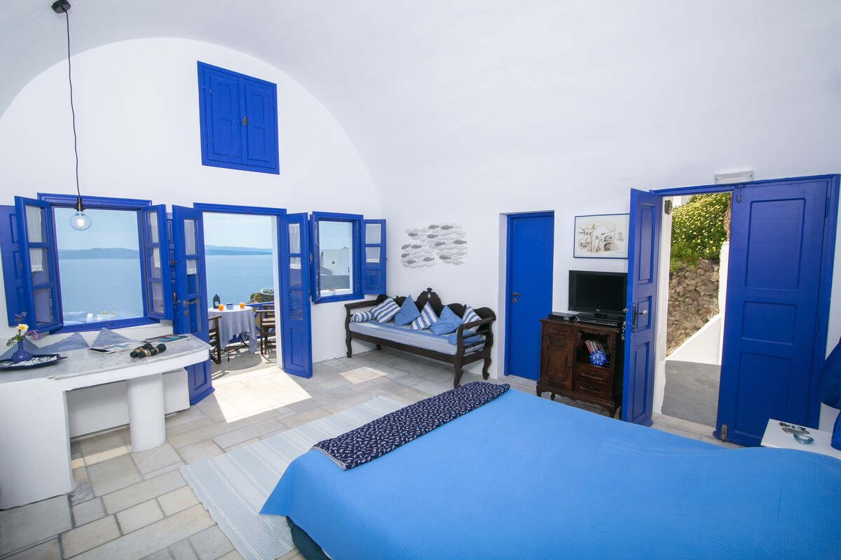 A spacious room features a king-size bed with a blue coverlet, complemented by light-colored tile flooring. Multiple large windows with blue shutters open to stunning views of the ocean. A seating area is present, along with a small dining space visible beyond the doorway.