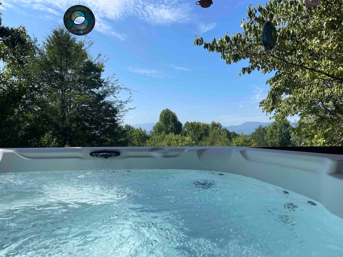 A brand new hot tub is shown, with water gently rippling under a clear blue sky. The surrounding greenery offers a serene backdrop, highlighting the distant mountains under bright sunlight. Decorative items hang above, adding a touch of charm to the outdoor setting.
