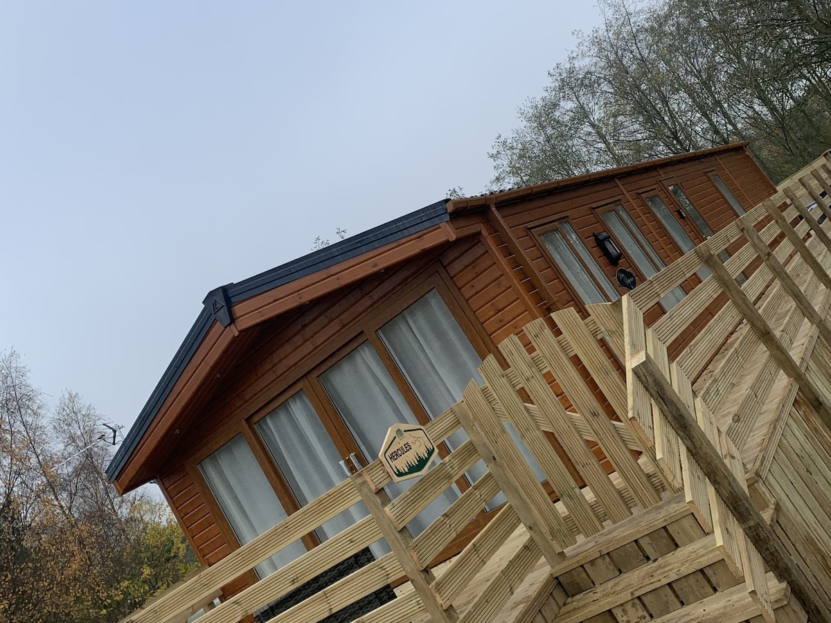The lodge features a wooden exterior with large windows offering natural light. A spacious wooden deck leads to the main entrance, enhancing accessibility. Surrounding trees are visible, contributing to the natural setting of the lodge.