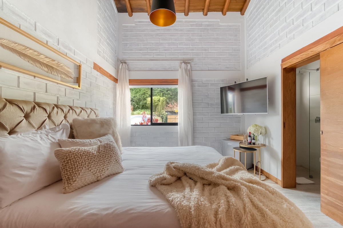 A comfortable bedroom features a large king-sized bed adorned with soft pillows and a cozy throw blanket. Natural light enters through a window, with light-colored curtains framing the view. A wall-mounted Smart TV is positioned across from the bed, and the bathroom entrance is visible.