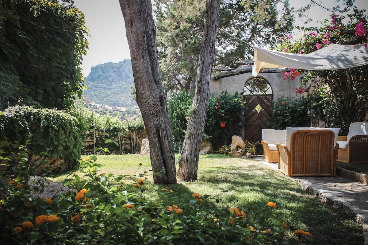 The garden features a well-maintained grassy area surrounded by lush greenery and colorful flowers. A shaded seating area with wicker chairs is visible, offering a serene view of the nearby hills. A decorative archway adds a charming focal point to the landscape.
