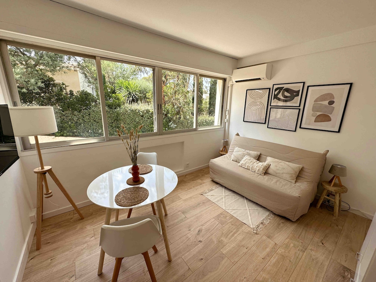 A bright living area features large windows that allow natural light to fill the space. A comfortable sofa is complemented by a dining table with four chairs. Artwork is displayed on the walls, enhancing the modern decor surrounded by greenery visible outside.