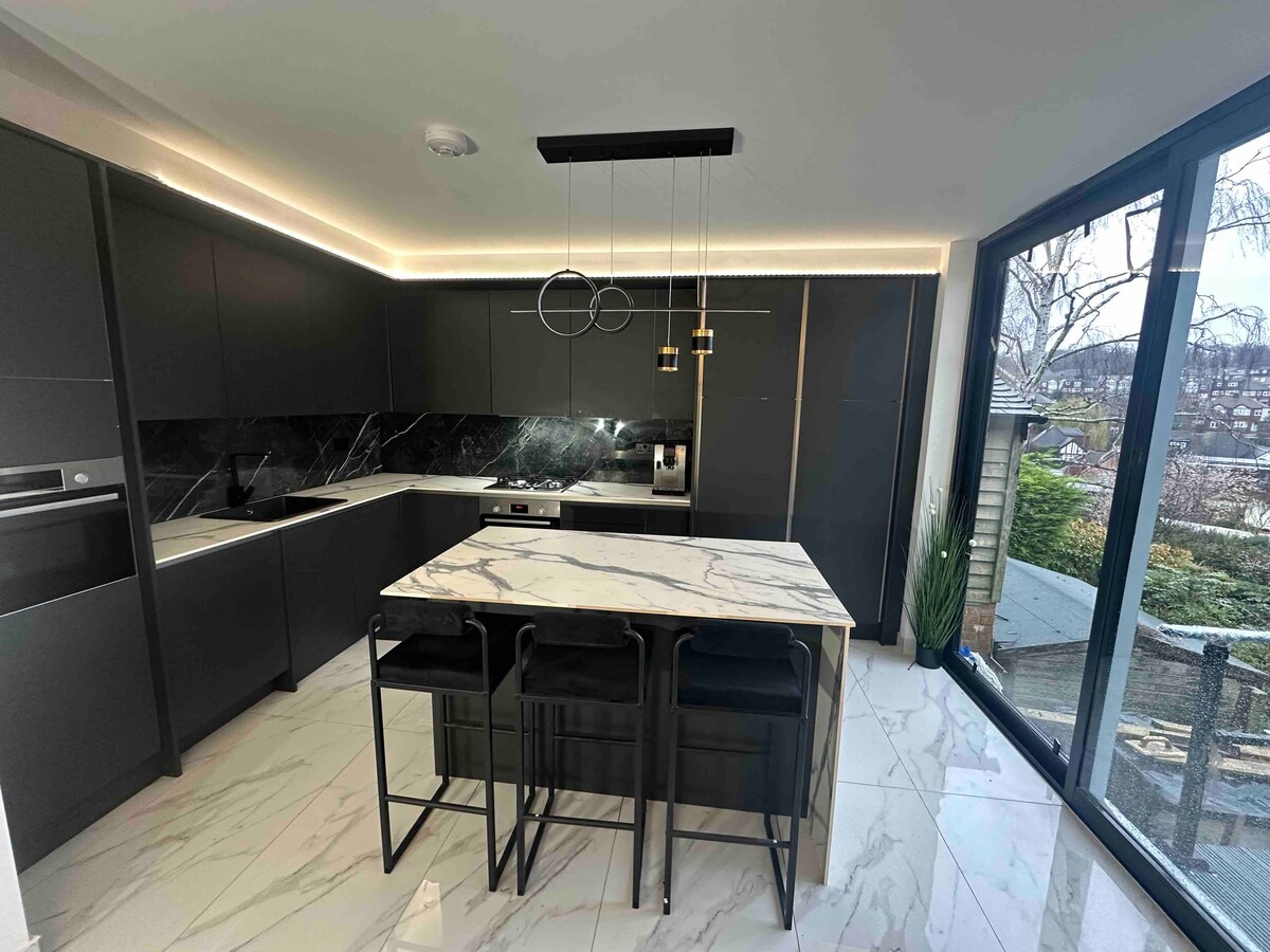 A modern kitchen features sleek black cabinetry and a spacious marble island with seating for three. Large windows allow natural light to flood the space, showcasing views of the garden outside. Stylish pendant lights illuminate the area, enhancing the contemporary design.