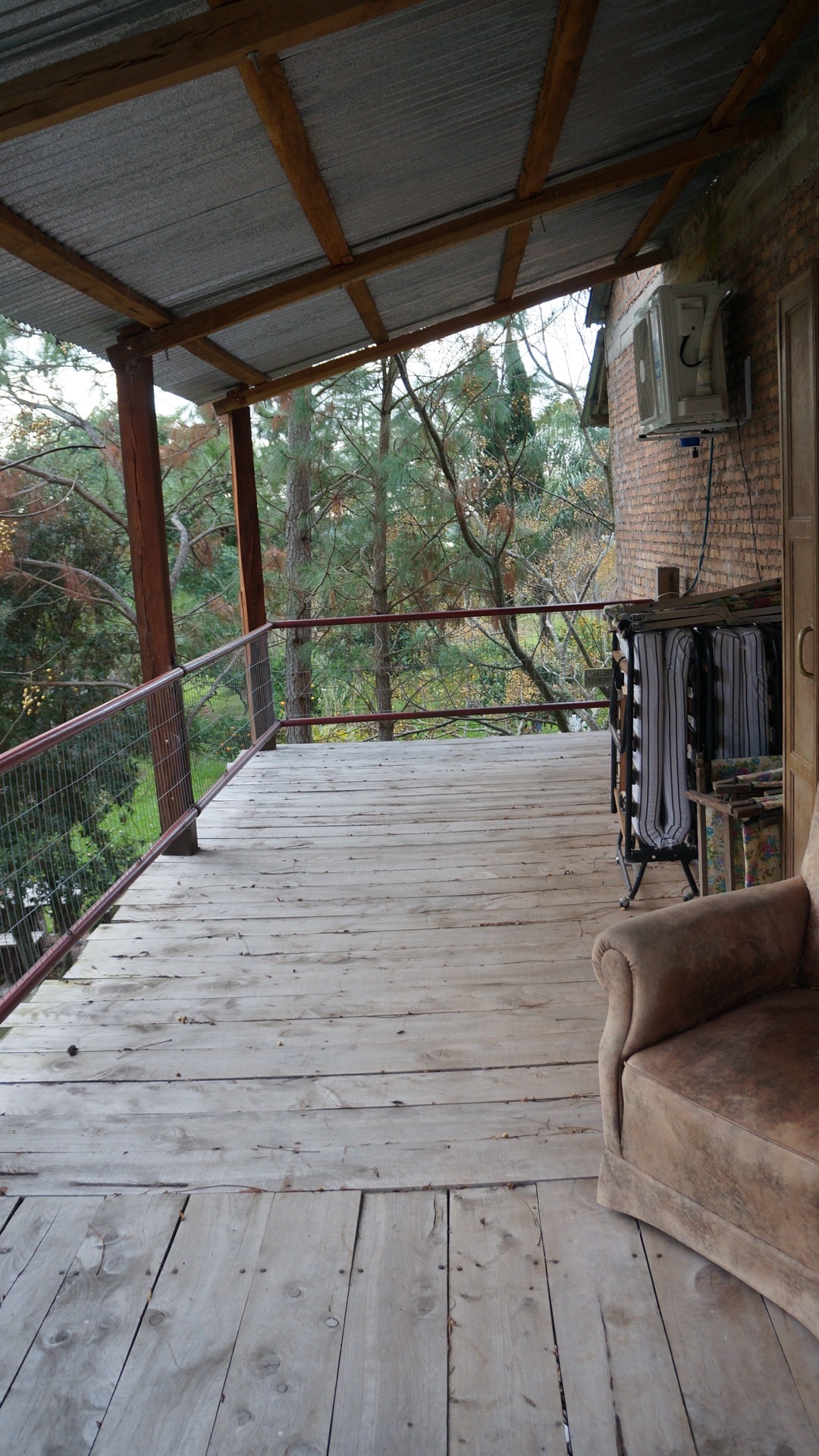A spacious wooden deck extends from the building, featuring a rustic railing that provides safety. A cozy armchair is positioned on the deck, while an air conditioning unit is mounted on the wall, offering a comfortable outdoor retreat surrounded by greenery.