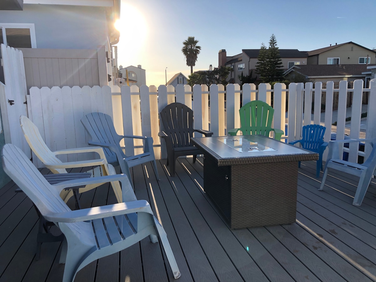 A spacious outdoor deck is surrounded by a white picket fence, featuring multiple Adirondack chairs in various colors and a central fire table. Sunlight casts warm hues, highlighting the inviting space ideal for relaxation and gatherings.