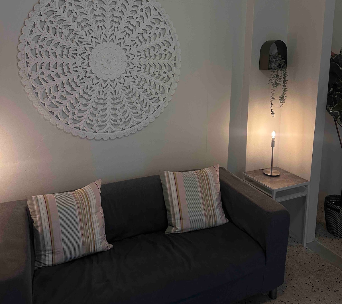 A cozy living area is highlighted by a plush gray sofa adorned with striped cushions. A delicate, circular wall art piece hangs above, while a soft glow is provided by a lamp and a candle on a side table, adding a touch of warmth to the space.