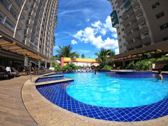 A large swimming pool is surrounded by tropical plants and lounge chairs. Bright blue water reflects the clear sky above. Two large buildings frame the space, enhancing the inviting atmosphere of the pool area.