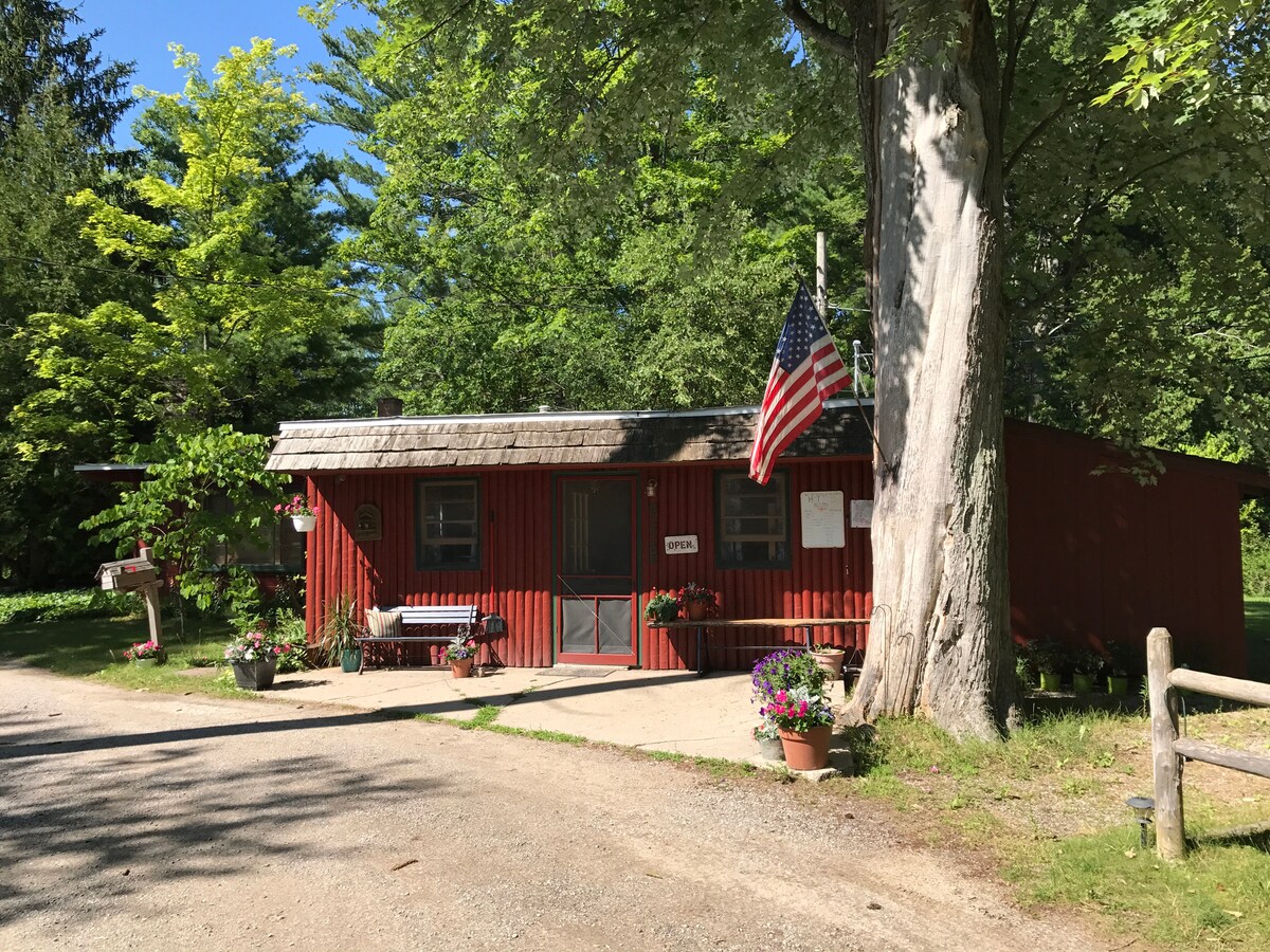 A charming log cabin exterior is surrounded by lush green trees and colorful flower pots. A decorative bench sits beside a welcoming entrance, while an American flag is proudly displayed. The gravel path leads to the door, enhancing the rustic setting.