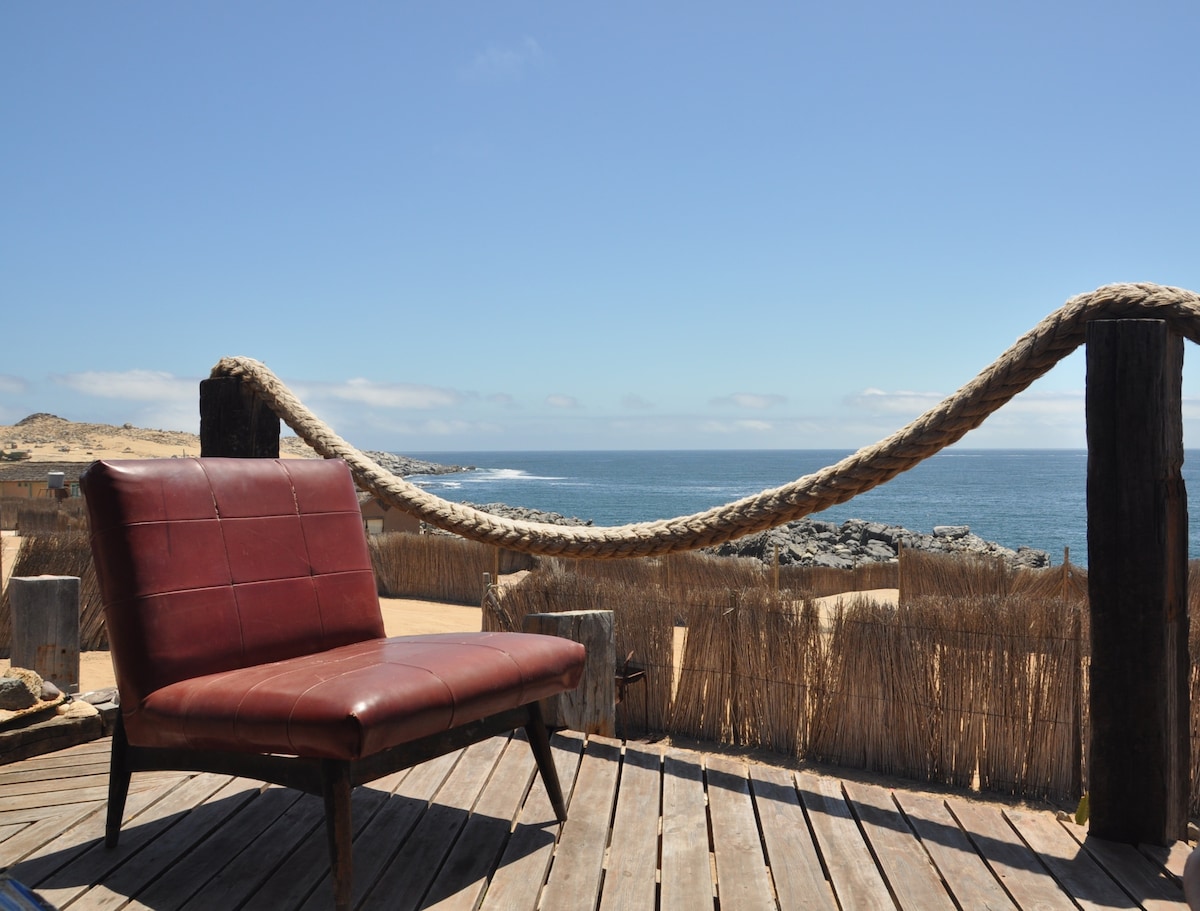 A wooden deck features a single brown lounge chair, facing the sea. Natural fibers are used in the surrounding fence and a thick rope runs along the edge, complementing the coastal view and clear blue sky.