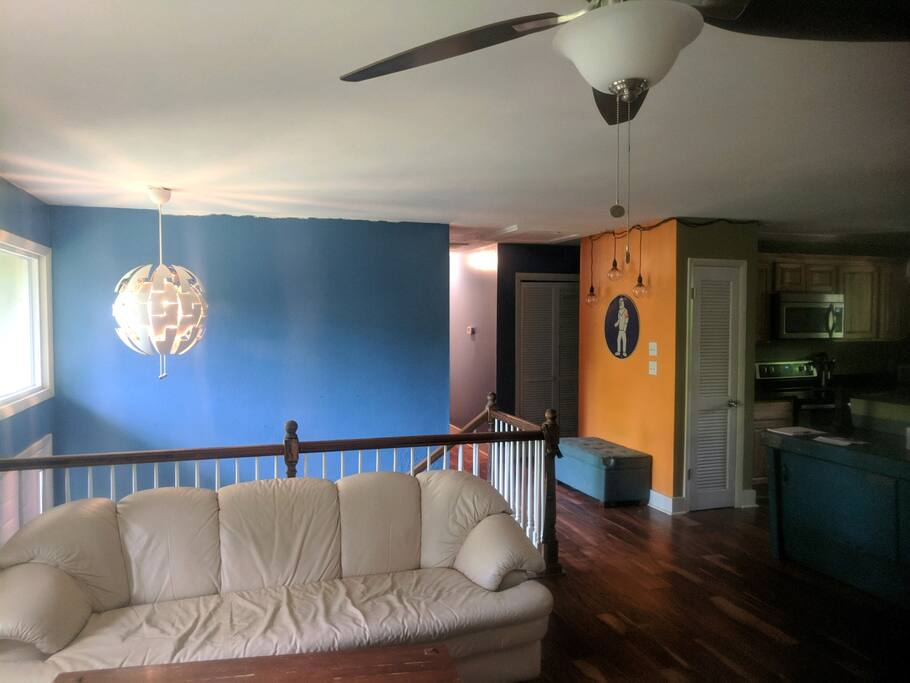 Large upstairs living area has two full size couches. Kitchen and dining is to the right and front door is down the half flight of stairs behind the couch. Hall closet is pictured with full bathroom just around the corner from the orange wall. 1st master bedroom is at the end of the hall.