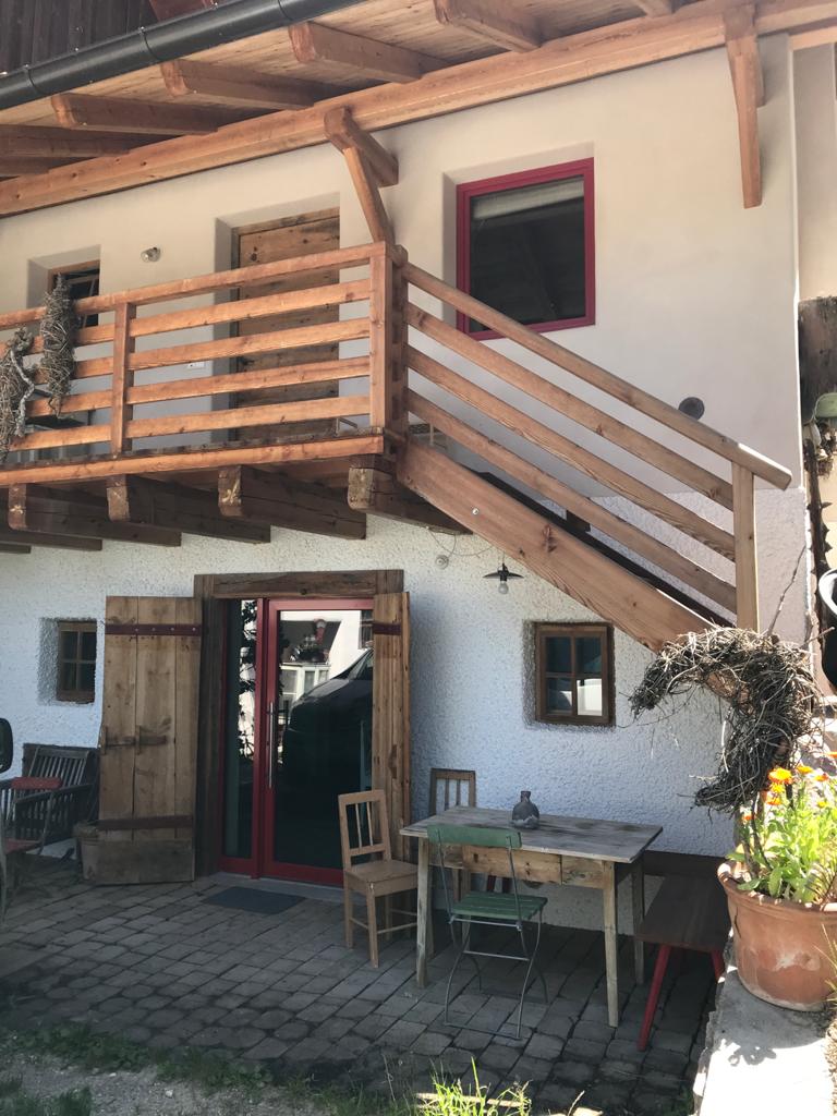The entrance of the studio features a wooden door framed by a rustic exterior. A small table and chair are positioned on the cobblestone patio, while a wooden balcony with sturdy railings extends above. Natural elements enhance the welcoming setting.