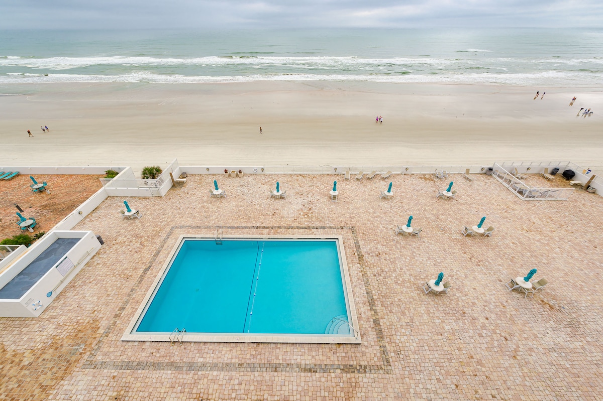 An aerial view captures a serene outdoor pool surrounded by a spacious deck, featuring numerous lounge chairs. The pool's clear blue water contrasts with the sandy beach and ocean waves in the background, creating a tranquil beachfront setting.
