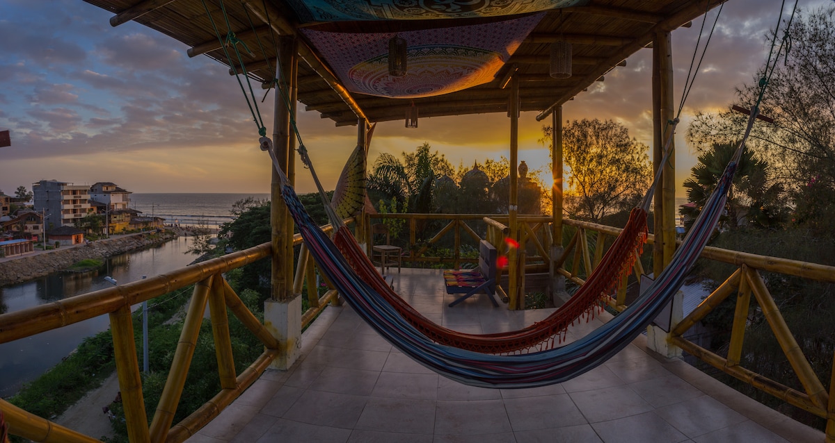 A spacious terrace is captured at sunset, adorned with a colorful hammock hanging from wooden beams. The ocean can be seen in the distance, while lush greenery surrounds the area. Soft lighting enhances the serene ambiance, inviting relaxation.