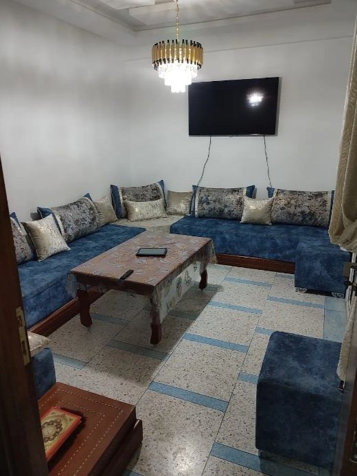 A cozy lounge area is furnished with a spacious blue seating arrangement, including cushions featuring floral patterns. A low wooden table is placed at the center, while a television mounts on the wall, and a chandelier offers ambient light from above.