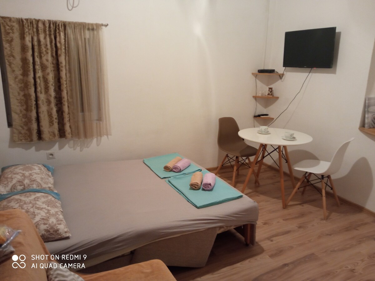 A cozy studio space features a spacious bed adorned with folded towels in various colors. A small dining area is equipped with a round table and two chairs. Natural light enters through a window dressed in light curtains, enhancing the bright atmosphere.