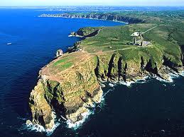 Aerial view captures a rugged coastline with steep cliffs bordering the deep blue sea. Green hills and patches of vegetation extend across the landscape, leading to a distant horizon where the land meets the water.