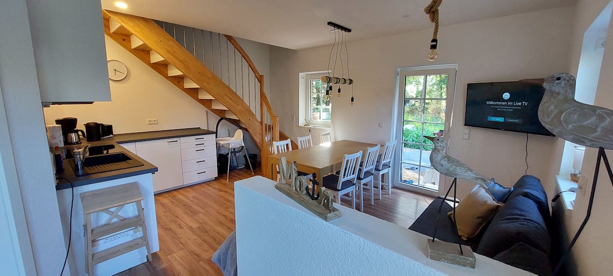 The open living area features a modern kitchen equipped with a coffee maker and dining table. A comfortable sofa faces a mounted TV, with a staircase leading to the upper level. Glass doors open to a view of the outdoor space, inviting natural light.