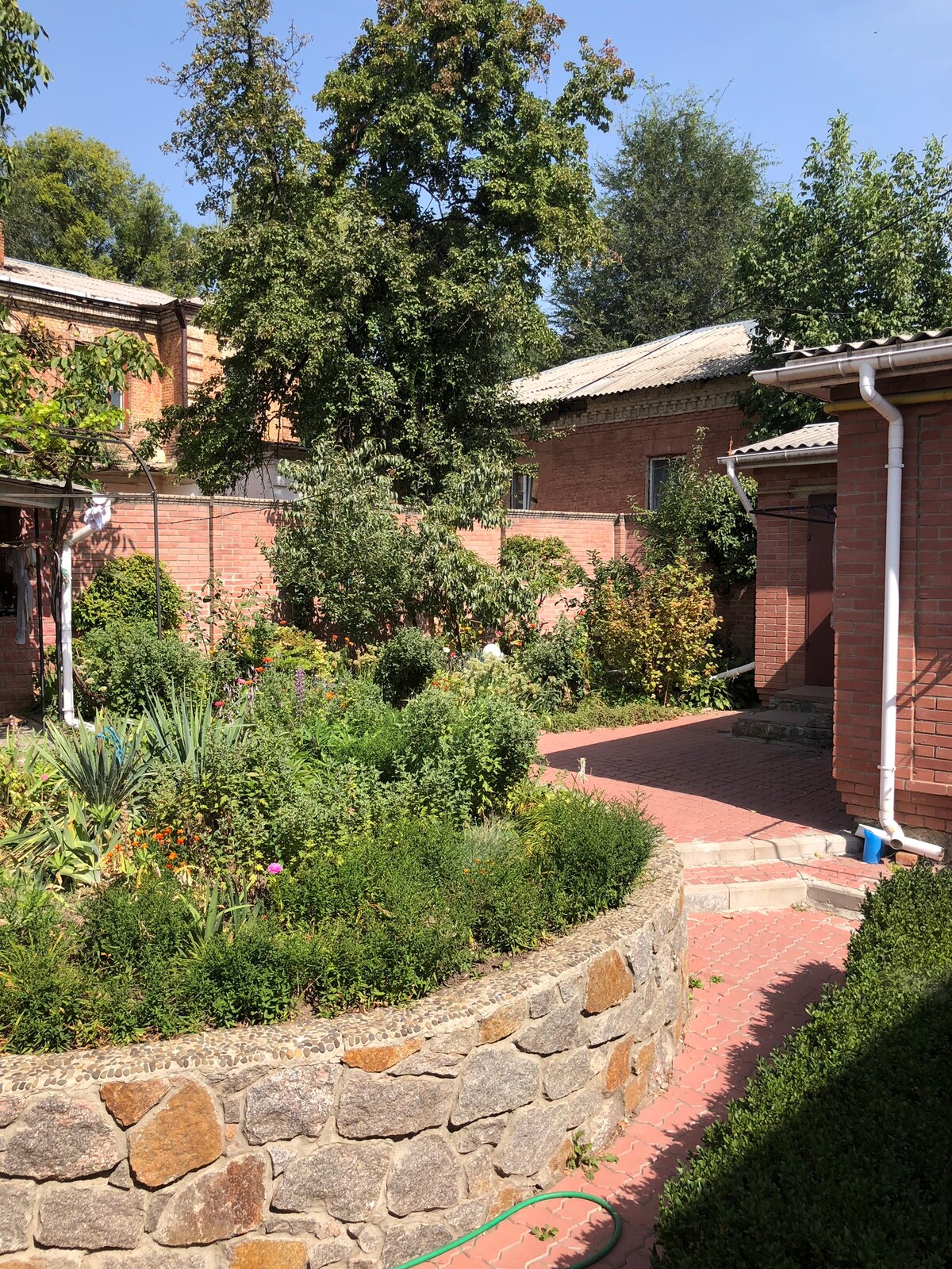 A peaceful yard is showcased, featuring a stone-bordered garden filled with various shrubs and plants. Green trees provide shade, and a brick pathway leads to a secluded area, enhancing the tranquil outdoor space.