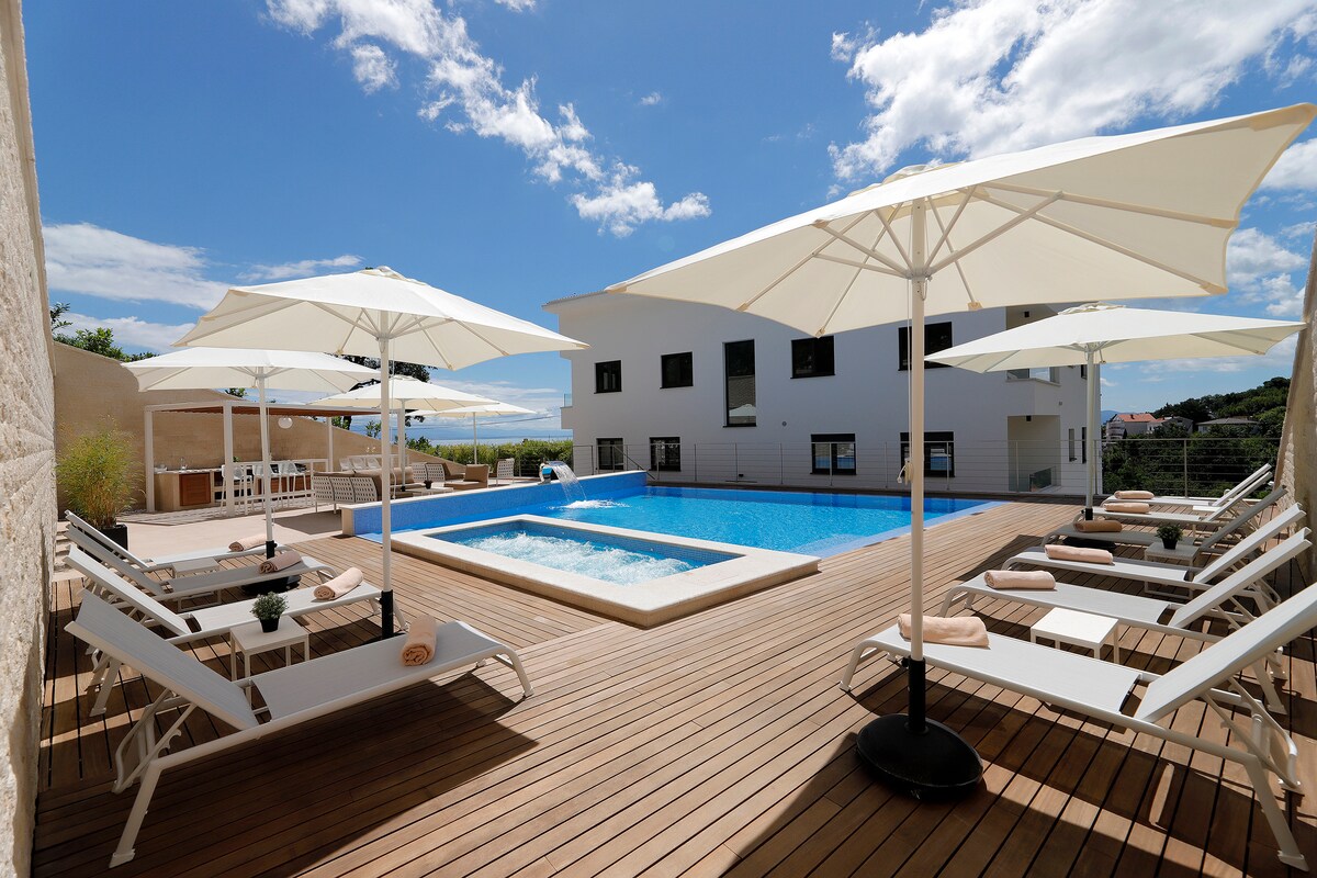 The outdoor area features a sun deck with several lounge chairs arranged around a heated pool and jacuzzi. Umbrellas provide shade over the seating area. A well-maintained garden and the building are visible in the background, under a clear blue sky.