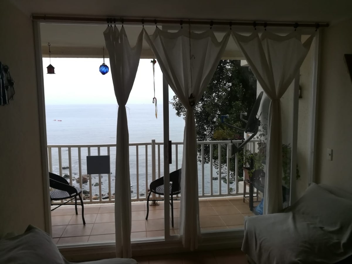 Floor-to-ceiling sliding glass doors offer views of the serene ocean. A small balcony is visible, furnished with two black chairs. Light curtains gently frame the opening, allowing natural light to fill the space while maintaining privacy.
