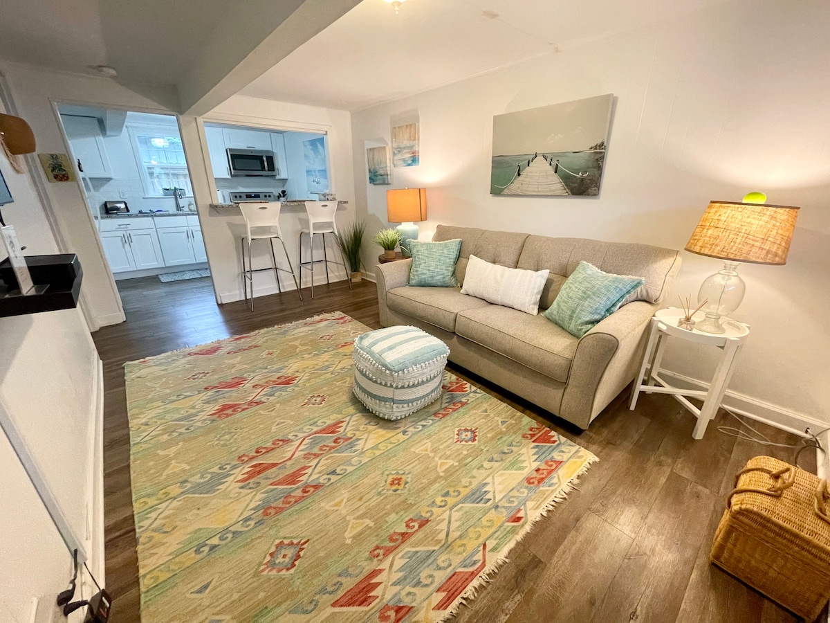 A spacious living area is depicted with a neutral-colored sofa adorned with decorative pillows. A patterned area rug lies on the floor, complementing the light wood tones. A small ottoman and a lamp add warmth to the space, while the kitchen is visible in the background.