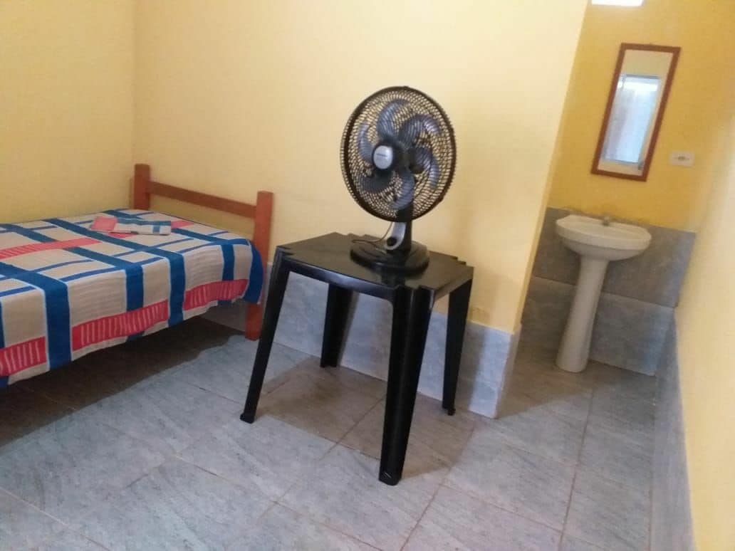 A simple room is presented with a wooden bed covered in a colorful patterned blanket. A black table holds a fan, and a small washbasin is positioned nearby, accented by a mirror. The walls are painted a light yellow, contributing to a bright space.