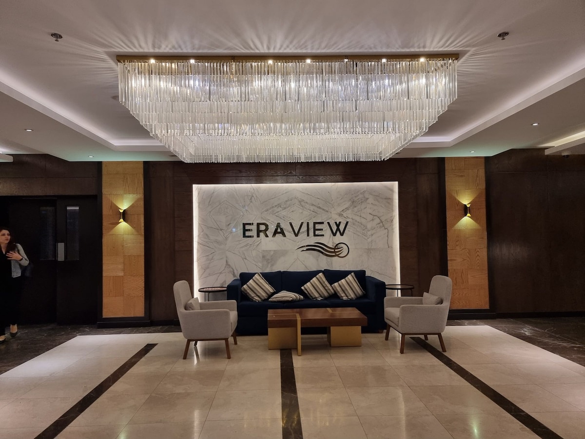 A modern lobby displays a large chandelier overhead, casting soft light across a polished marble floor. The backdrop features the 'ERAVIEW' logo against a marble wall, while two stylish armchairs with decorative cushions are positioned around a wooden coffee table.