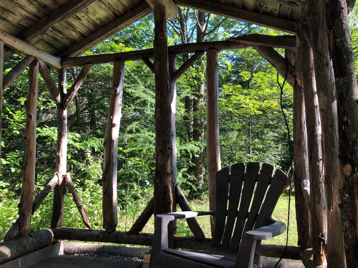 A rustic outdoor seating area features a large wooden chair framed by natural timber supports. Lush green trees are visible in the background, creating a serene and shaded environment.