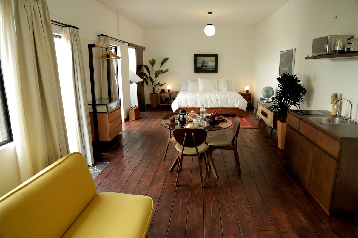 A spacious loft environment is presented with a bright bed area featuring multiple pillows. A dining table is set for two, and a compact kitchen unit is visible. Sunlight enters through large windows, illuminating the warm wooden flooring and surrounding greenery.