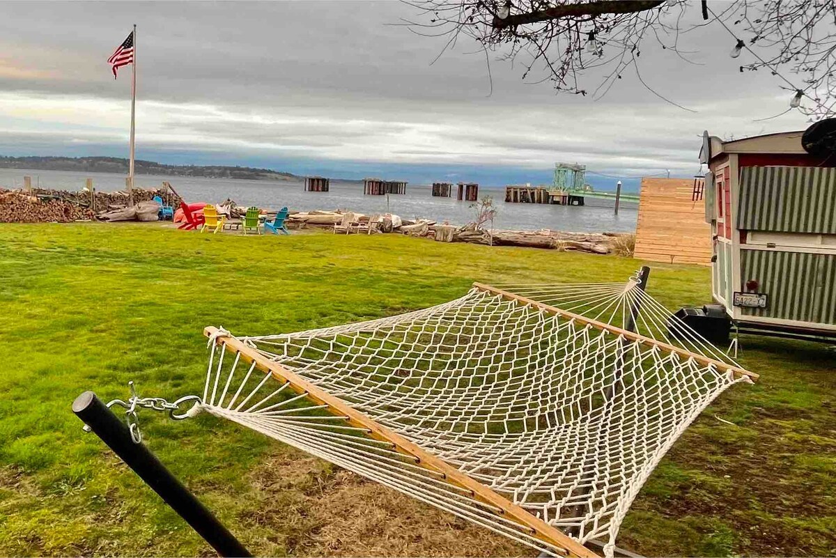 A hammock is positioned on a grassy area, offering a comfortable resting space with views of the waterfront. Colorful Adirondack chairs are arranged nearby, and a rustic wooden dock extends into the water, creating a tranquil coastal atmosphere.