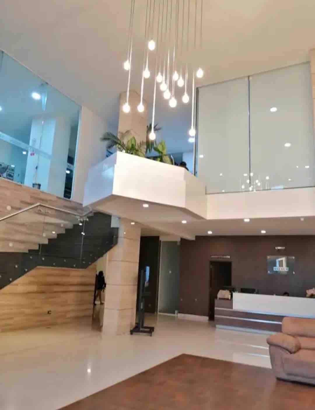 A spacious lobby is featured with high ceilings and large windows allowing natural light to fill the area. Modern pendant lights hang above the sleek staircase, while greenery adds a touch of freshness. A reception area is visible in the background, contributing to an inviting environment.