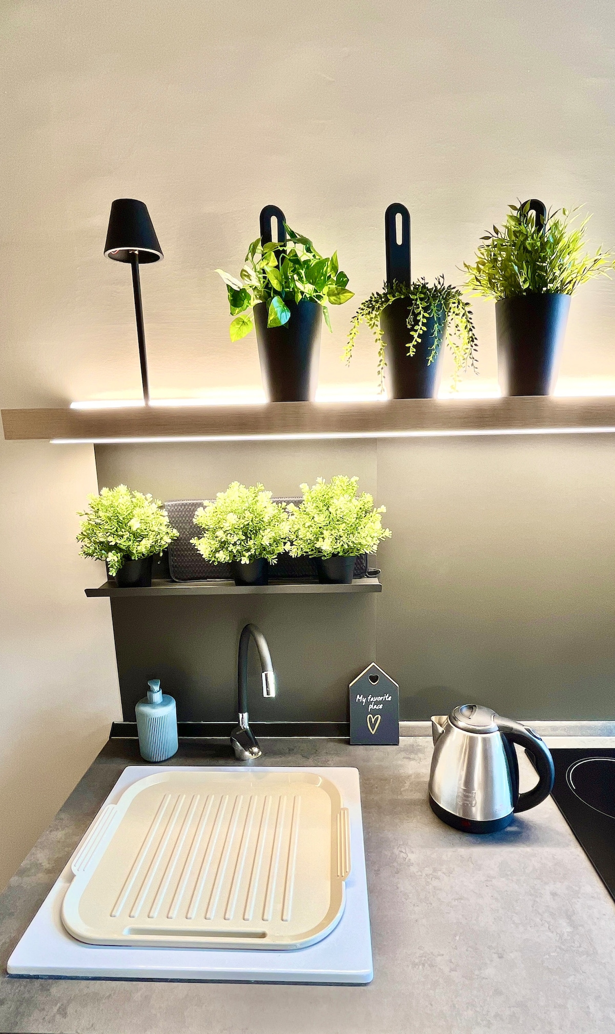 A modern kitchen setup features a countertop with a drying rack and a sleek kettle. Above, a shelf displays several potted plants, including leafy greens and herbs, complemented by a warm light strip. A soap dispenser is positioned nearby, enhancing functionality.