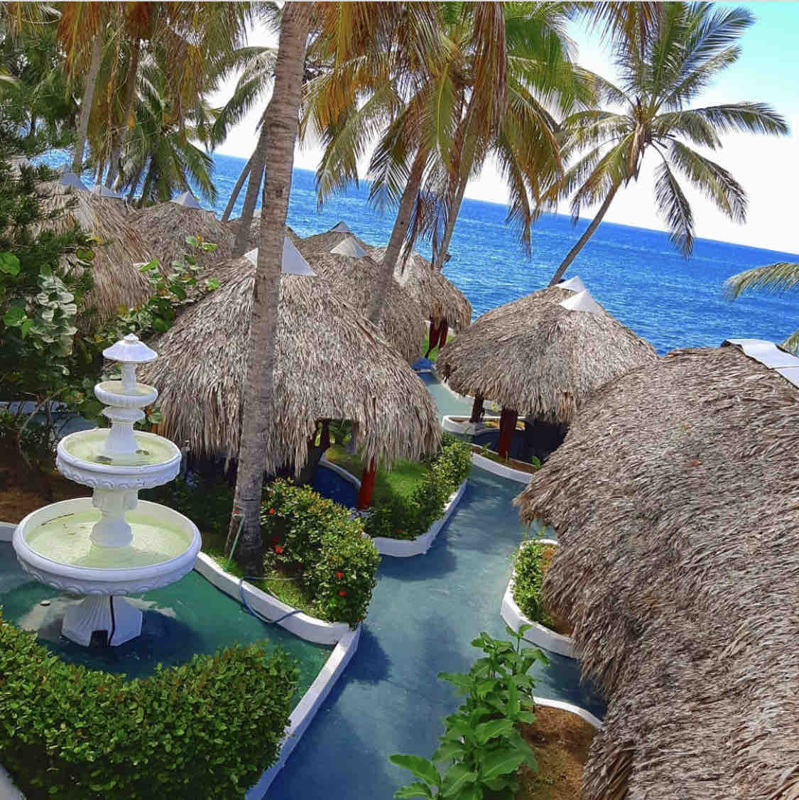 Tropical palm trees surround thatched-roof cabanas, creating a serene outdoor space. A decorative fountain is positioned at the center, adding to the calm ambiance. The blue ocean is visible in the background, enhancing the peaceful atmosphere of the area.