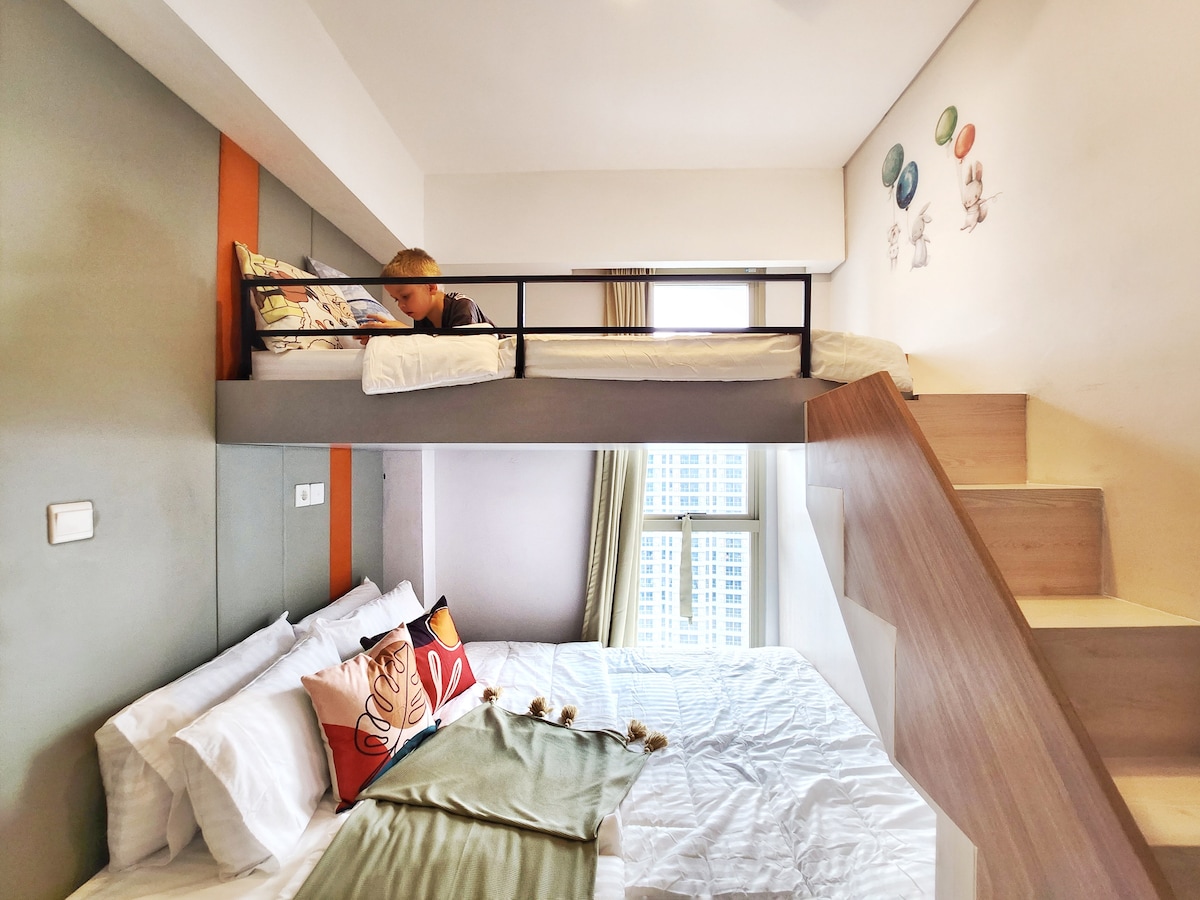 A cozy sleeping area is showcased with two bunk beds, designed for comfort. A spacious bed with soft linens is placed below, while a single bed lofted above allows for additional sleeping space. Light filters through a nearby window, enhancing the inviting ambience.