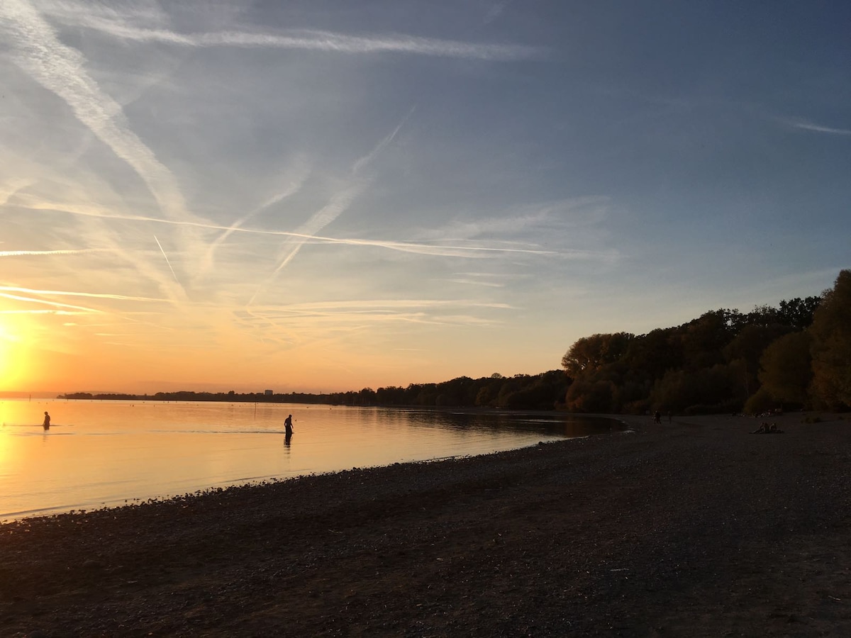The image captures a serene lakeside view during sunset. A calm body of water reflects the warm colors of the sky, with silhouettes of people enjoying the tranquil environment. The shoreline features pebbles and trees, providing a peaceful natural setting.