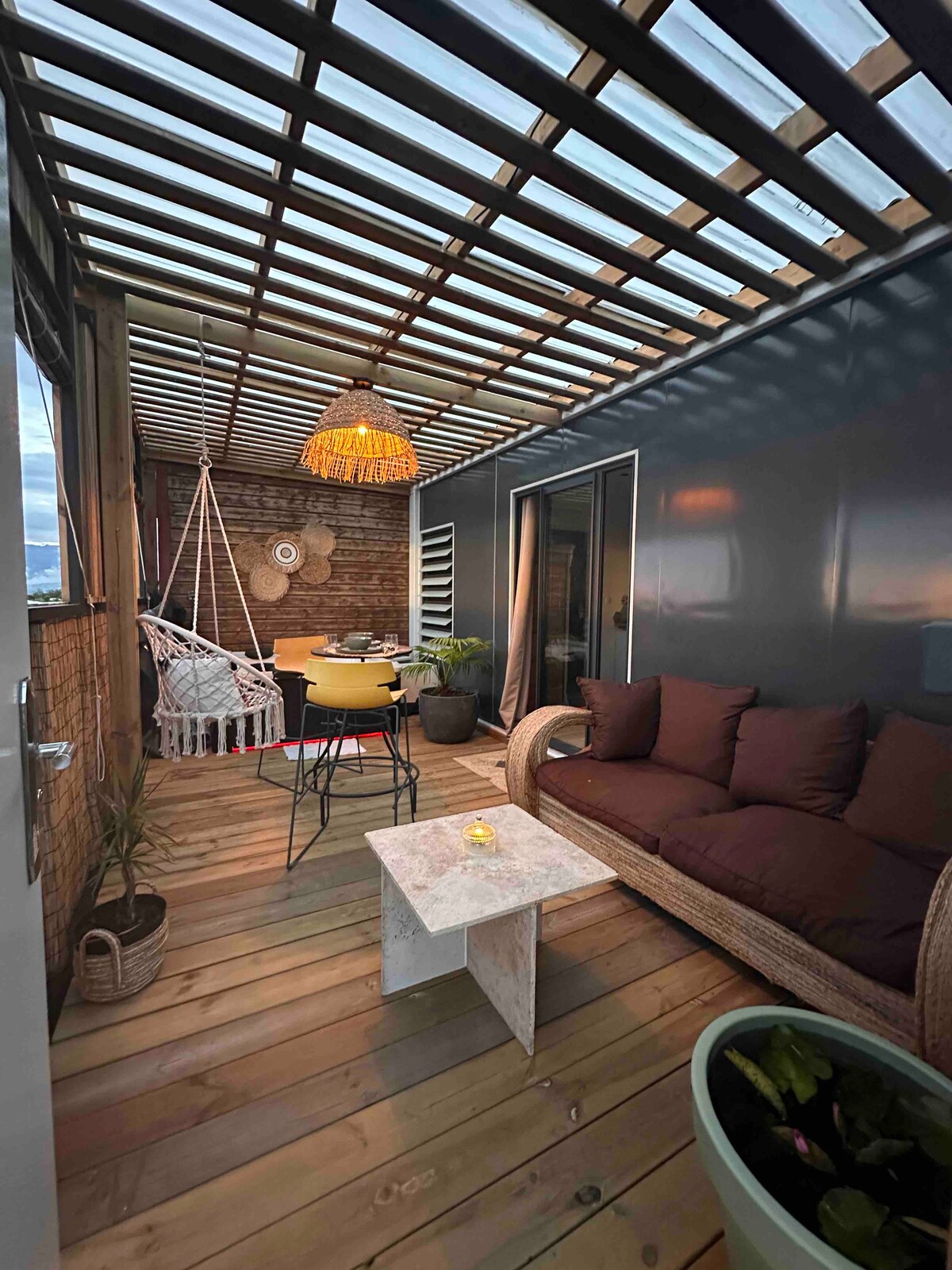 A covered terrace features a comfortable sofa with dark cushions, surrounded by wooden flooring and potted plants. A round dining table with chairs is set for outdoor dining, while a hanging chair adds a relaxing touch to the space, illuminated softly by a pendant light.