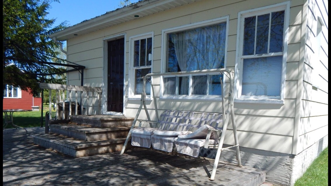 A porch area is shown, featuring a white swing set, providing a comfortable seating option. The entrance is marked by a dark door, flanked by large windows that allow ample natural light. The surrounding grass is well-maintained, contributing to a serene outdoor environment.