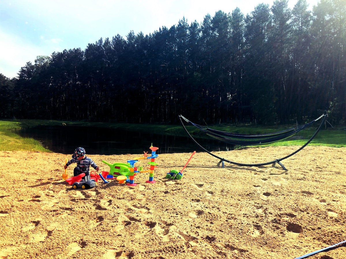 A sandy area by a serene pond is depicted, featuring colorful children's toys. A child is playing with a small vehicle and building blocks near a softly swaying hammock set up nearby. Lush trees line the background, enhancing the natural setting.