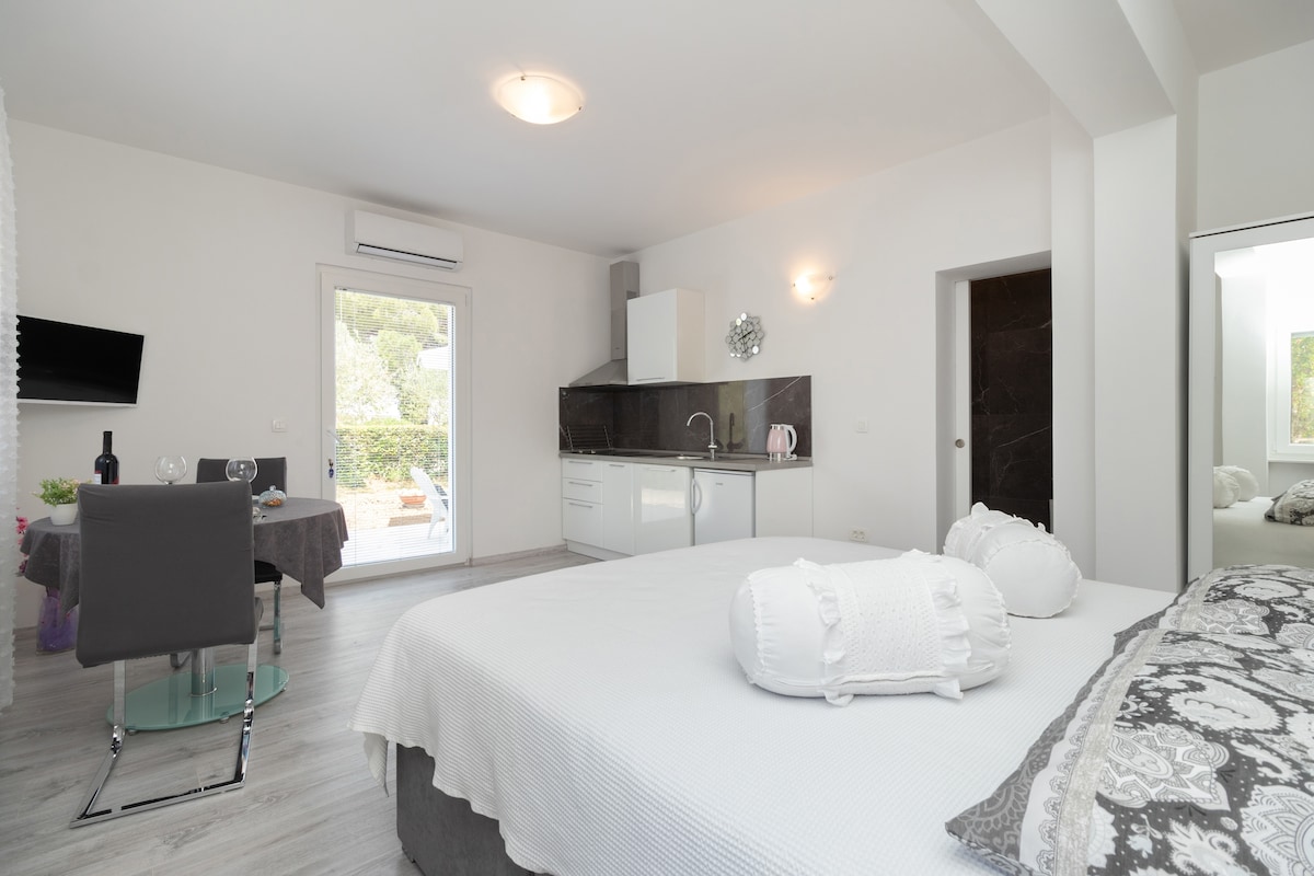 A modern studio apartment is showcased, featuring a spacious bed with decorative pillows. The kitchenette is equipped with essential appliances. A dining table set for two is visible, alongside a wall-mounted TV. Natural light enters through the sliding glass doors leading to an outdoor area.