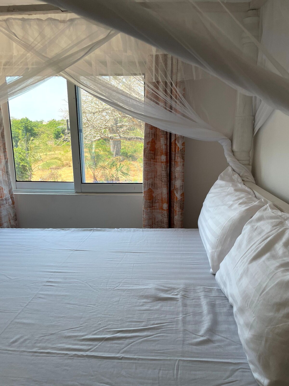 A cozy sleeping area features a spacious bed with crisp white linens. Sheer drapes frame the large window, allowing natural light to illuminate the room. Views of nature are visible through the window, enhancing the calming atmosphere.