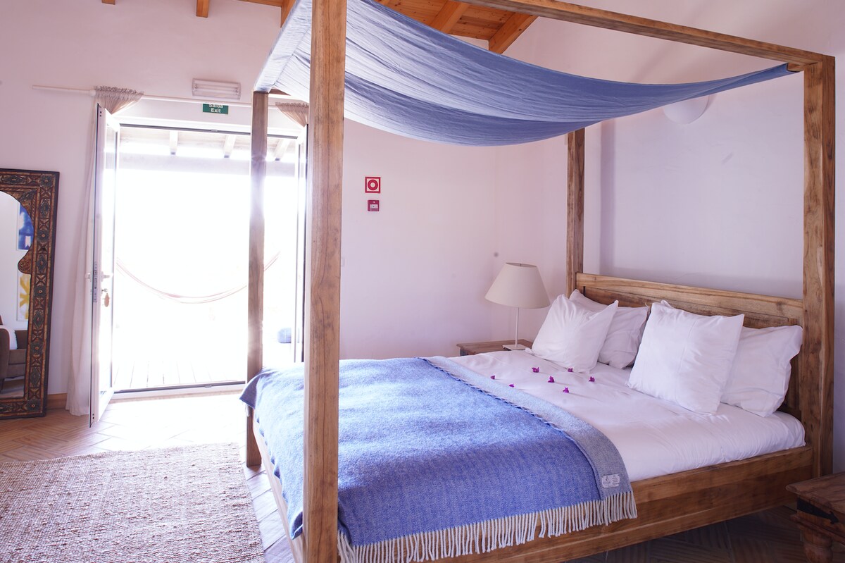 A spacious room features a wooden four-poster bed adorned with white bedding. A blue blanket drapes across the bed, while a light fixture illuminates the space. A doorway leads to a balcony area with a hammock, allowing natural light to fill the room.