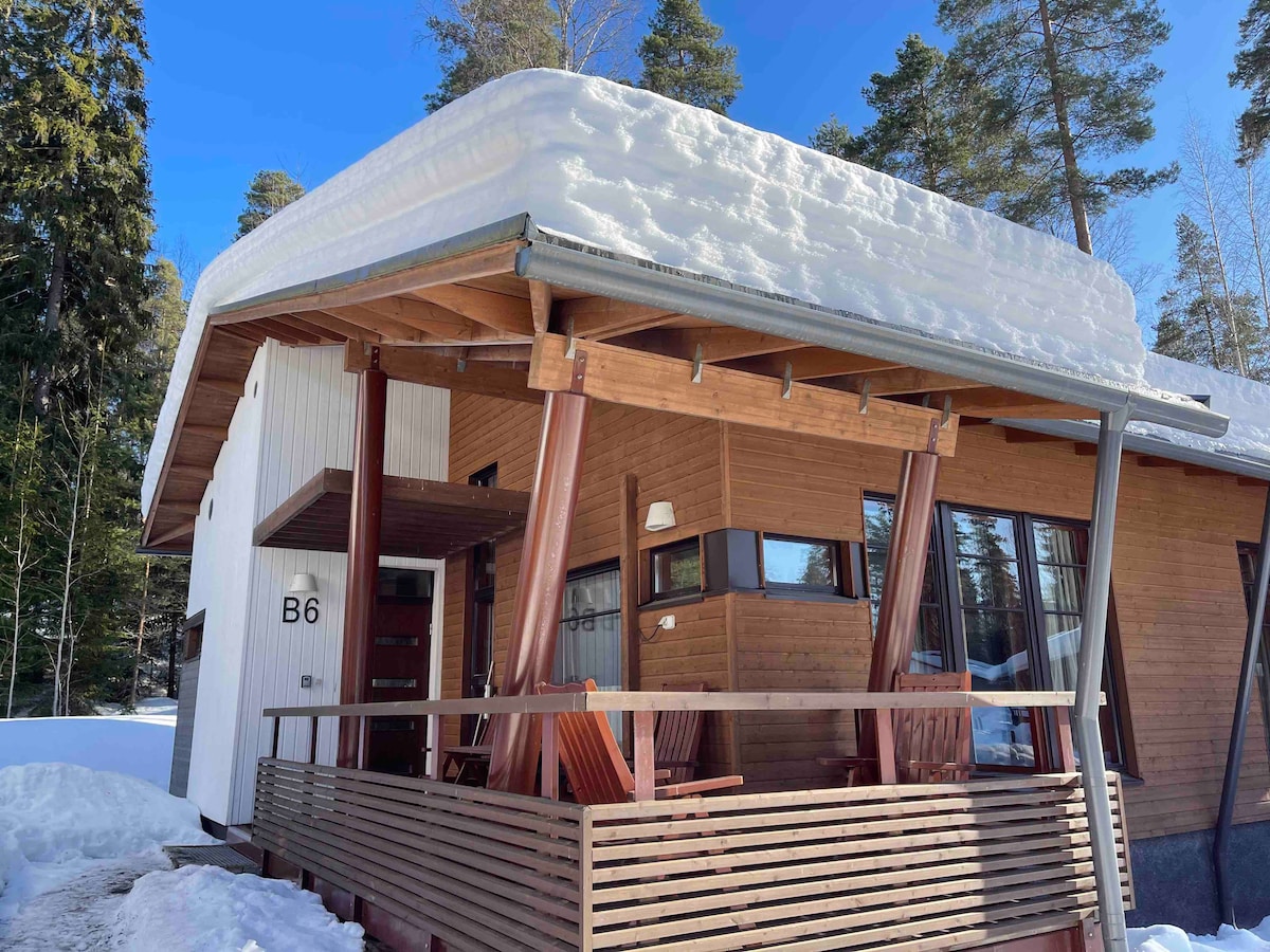 A modern wooden cottage is framed by a snowy landscape, showcasing a sloped roof covered in a layer of snow. The entrance is highlighted by large windows and a wooden deck with seating, surrounded by tall trees under a clear blue sky.